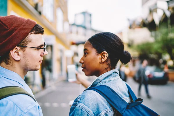 Cropped back view of african american young woman together with caucasian boyfriend looking at each other standing in urban setting and making selfie photo on modern smartphone device