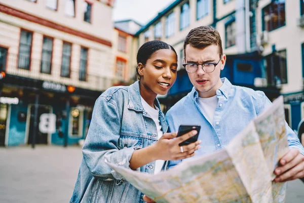 忧心忡忡的高加索年轻人拿着旅行地图 与非洲裔美国人女友一起寻找路径 在城市的城市环境中 他们用在线导航软件展示智能手机 — 图库照片