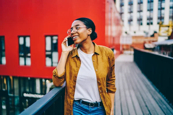 Smiling dark skinned hipster girl in spectacles satisfied with tariffs for mobile calls in roaming talking on cellphone, positive african american woman having conversation on smartphone on stree