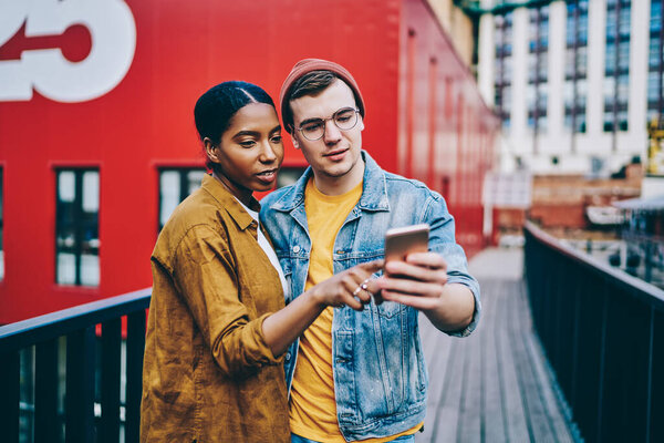 Multiracial couple in love dressed in trendy wear checking new app for searching places of interest in city on cellphone, male and female millennial hipsters read news from social network on smartphon