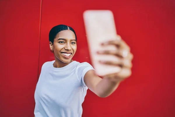 Cheerful dark skinned young woman posing for selfie on modern smartphone camera standing on red wall background, happy dark skinned hipster girl holding mobile phone taking picture via applicatio