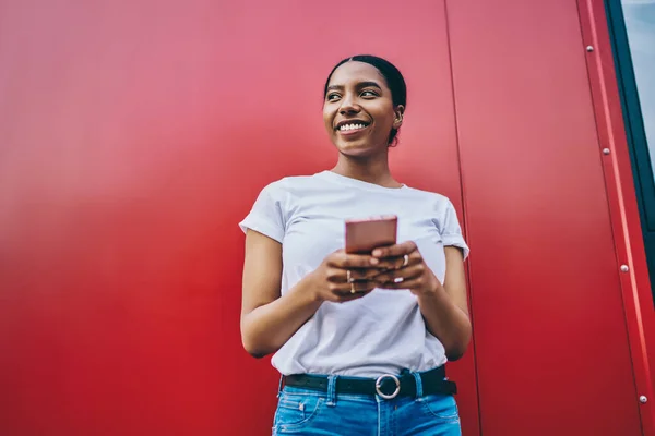 Smiling young african american woman in shirt with copy space for brand name or label holding mobile phone and looking away, cheerful dark skinned female blogger using smartphone stand on red wall