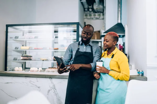 アフリカ系アメリカ人男性と女性のエプロンは熱い飲み物を楽しみながら カフェで休憩中にカウンターの近くに立っている間 モダンなタブレットを使用しています — ストック写真