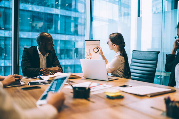 Young elegant woman sitting at table with multiracial coworkers in office boardroom showing paper and doing presentation about statistical data