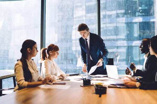 Young formal man showing paper document to group of multiethnic coworkers sitting together at table in sunny boardroom of office