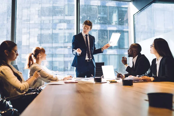 Young formal man with document standing at table in front of multiracial colleagues doing presentation in boardroom of modern office