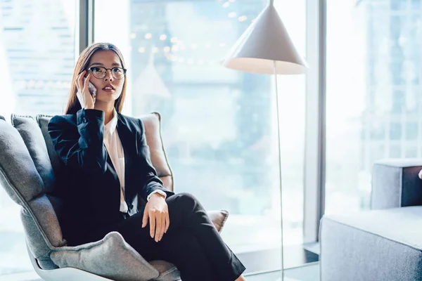 Young trendy Asian woman in suit and glasses sitting in armchair and having phone call on smartphone in contemporary office