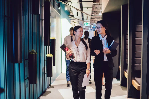 Young elegant diverse women carrying files and smartphones while walking together in contemporary office hallway and chatting friendly having discussion
