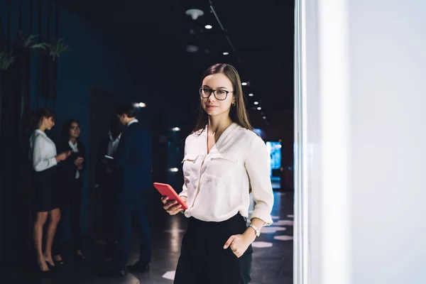 Beautiful and confident woman in glasses and elegant outfit holding mobile phone and looking at camera standing in office hall
