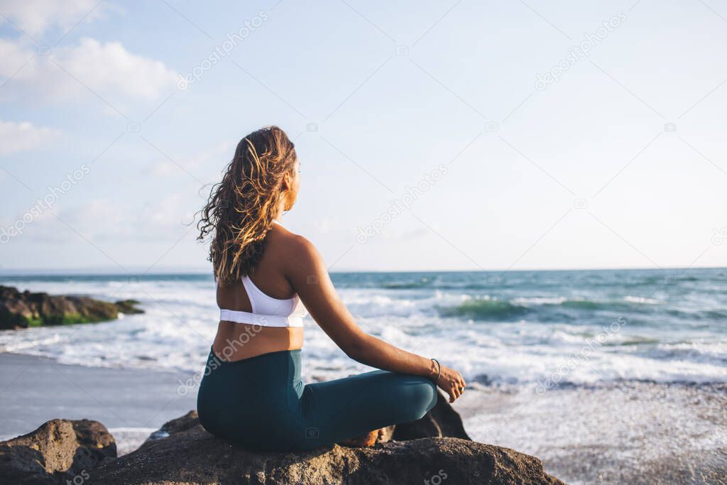 Yoga on beautiful Indian beach performed by attractive young African American girl dressed in sport tracksuit, evening spiritual training near ocean by calm female in lotus pose meditating