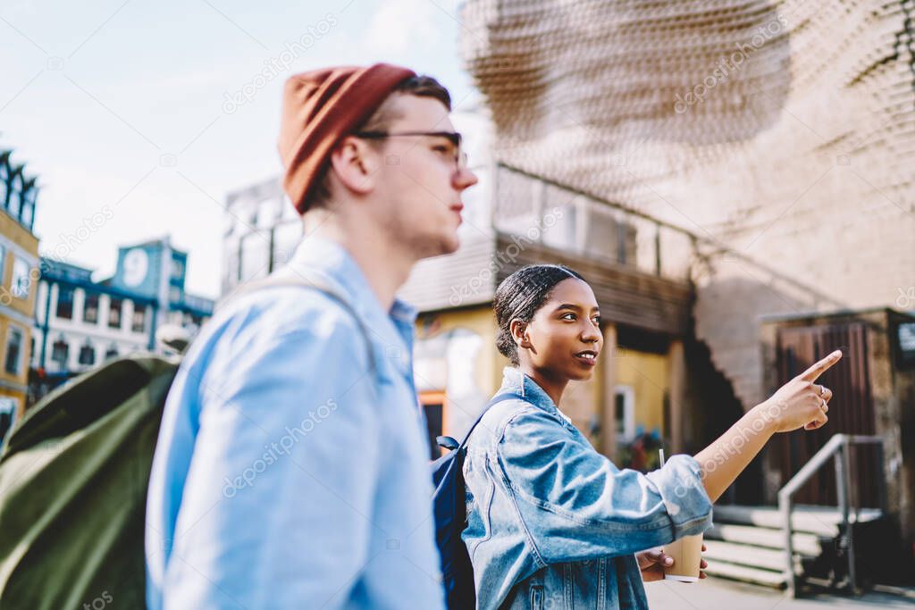 African american young woman pointing right direction while walking in urban setting together with hipster guy during weekend's trip.Diverse couple in love dressed in casual wear talking about route