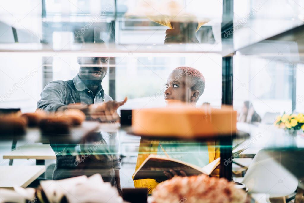 African American man and woman pointing at cake display and writing down dessert assortment into open notebook while working in bakery together