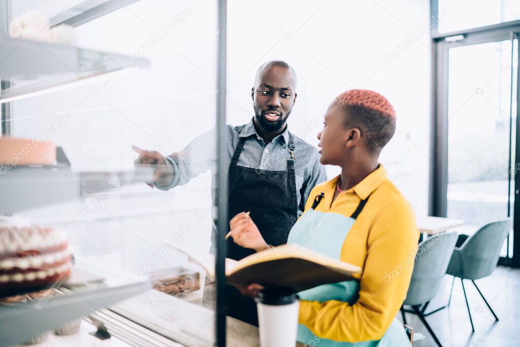 Black man in apron pointing at cake display and looking at waitress with open notepad while working in small cafe and making accounting records