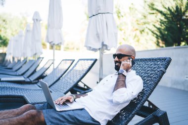 Beyaz gömlekli ve güneş gözlüklü yakışıklı adam terasta şezlongda uzanmış cep telefonuyla konuşurken dizüstü bilgisayarla çalışıyor. 