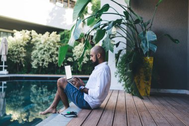 Gömlekli ve şortlu bir adamın bilardonun kenarında oturup uzaktan kumandayla tatil yapması. 