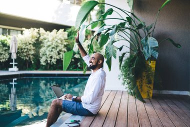 Havuzun kenarında oturmuş, iş gezisi yaparken el sallayan neşeli etnik adamın yan görüntüsü. 