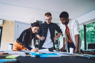Genç çoklu etnik çeşitliliğe sahip kadın ve erkekler ofiste birlikte duruyor ve yeni bir plan oluştururken kağıda dökülüyorlar. 