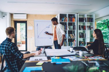Sıradan bir Afro-Amerikalı adam, masada oturan genç erkek ve kadına sunum yaparken güverteye grafik çiziyor. 