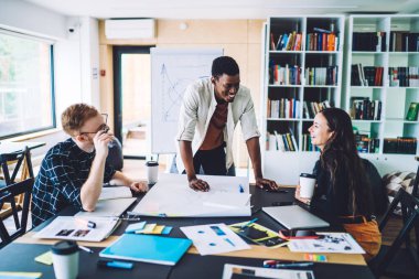 Elinde kahve fincanı olan çok ırklı genç erkek ve kadın, modern ofiste masasında oturmuş, kahkahalar atıyorlar. 