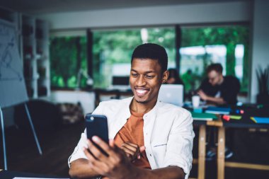 Çağdaş ofiste, arka planda meslektaşlarıyla masada oturan neşeli, sıradan siyah bir adam ve gülümseyen modern akıllı telefon kullanıyor. 