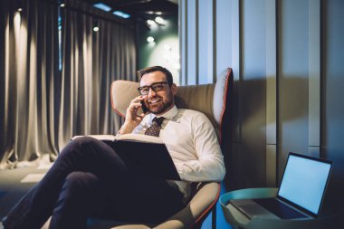 İş ortağıyla mobil sohbet sırasında koltukta oturan ve kameraya gülümseyen yakışıklı erkek tüccarın portresi, laptop cihazıyla kopyalama alanı ile eşzamanlı çalışma alanı