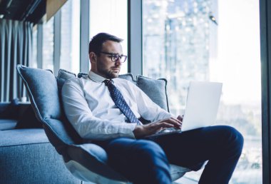 Gözlüklü yakışıklı adam dijital teknolojiyle online muhasebe yapıyor. Ofis kablosuz ağına bağlı. Modern dizüstü bilgisayarla belgelerle çalışan zeki bir erkek tüccar.