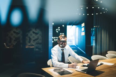 Optik gözlüklü mutlu işadamı, çalışma masasında oturup kağıt belgelerini analiz ediyor, bilgisayarında dijital iş yönetimi ve ortakların iletişimini inceliyor.