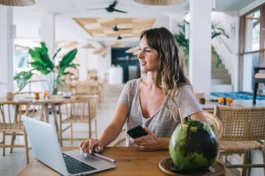 Kafkasyalı serbest çalışan kadın Endonezya tatillerinde internet üzerinden dağıtım olanaklarını kontrol ediyor. Kafeterya masasında dizüstü bilgisayar ve egzotik hindistancevizi için oturan çekici kadın.