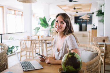 Modern cep telefonu ve dizüstü bilgisayarlı pozitif Kafkas dijital göçebelerinin portresi Bali kafeteryasında hindistan cevizi içecekleriyle iş için zaman harcıyor, kameraya gülümseyen mutlu serbest çalışan