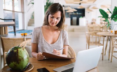 Ciddi bir Kafkas Endonezya kafeteryası yöneticisi. Laptop aygıtı ve tropikal meyve ile masada çalışırken, kağıt ders kitabından gelen teslimat bilgileri hakkında notları kontrol ediyor.
