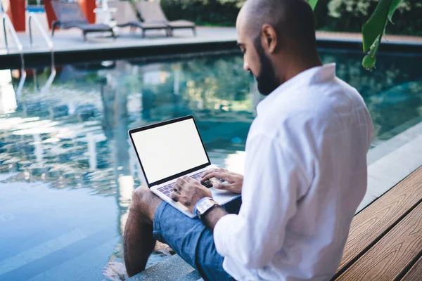 Side view of lounging ethnic men refreshing feet in cool water of resort pool while sitting on edge and using laptop