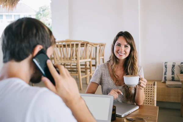 快乐微笑的女人的画像 一杯卡布奇诺在镜头前微笑 等待忙碌的丈夫打完智能手机 看到熟练的男人在空白的笔记本电脑上工作 通过手机交谈 — 图库照片