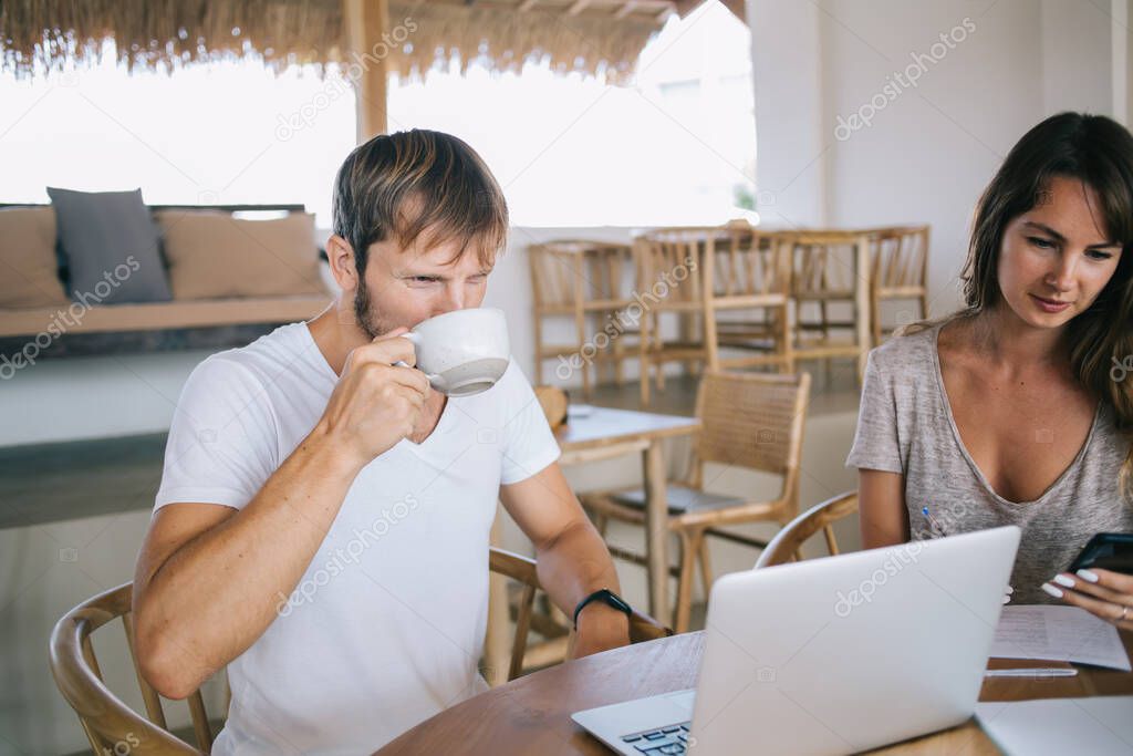 Skilled hipster blogger 30s drinking coffee and watching online webinar about personal growth while Caucasian wife checking mail via smartphone gadget using wireless wifi in cafeteria on Bali