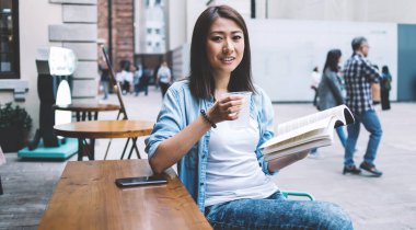 İlginç çok satan kitapların edebiyat alanındaki zeki kadın portresi, elinde kitap ve su bardağıyla şehir merkezinde kameraya bakan gülümseyen genç bir kadın.