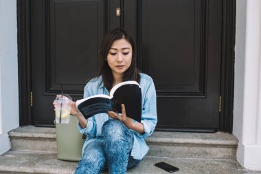 Çinli kız öğrenci elinde soğuk su içeceği tutan ilginç edebiyat kitapları okumaya odaklanmış güzel genç bir kadın boş zamanlarını şehrin merdivenlerinde en çok satanlar listesinde dinlenerek geçiriyor.