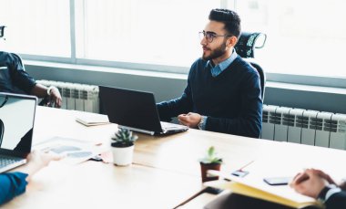 Optik gözlüklü kendine güvenen genç bir erkek yönetici yeni modern bir dizüstü bilgisayarla ofiste oturuyor ve çalışma saatlerinde iş arkadaşlarıyla bilişim projesiyle ilgili fikirleri tartışıyor.