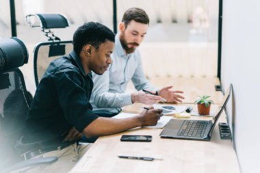 Dizüstü bilgisayarda iş uygulaması oluşturma projesini tartışan iki profesyonel erkek, şirket için muhasebe alışverişini analiz eden çok kültürlü genç meslektaşlar