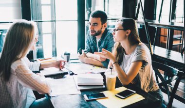 Kafede toplanan genç kadın ve erkeğin yan görüntüsü. İçki içip, not defterleri ve kağıtlarla sohbet ediyorlar.