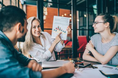 Bir grup mutlu genç kadın ve erkek kafeteryada oturmuş renkli grafiklerle kağıtları tartışıyor ve fikirlerle paylaşıyorlar.