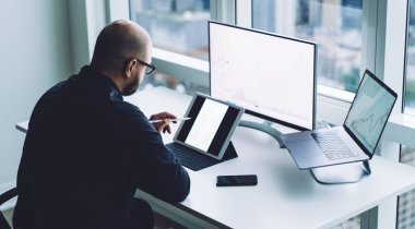 Büyük pencereli çağdaş ofiste dizüstü bilgisayar ve bilgisayarla oturup tablet izleyen gözlüklü kel işadamının yan görüntüsü