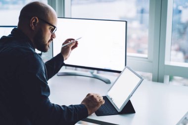 Modern ofiste pencereye karşı bilgisayarlı tablet kullanan gözlüklü yetişkin sakallı adamın yan görüntüsü