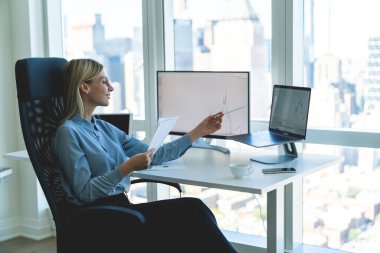 Modern ofisteki bilgisayar masasında çalışan güzel, kendine güvenen bir iş kadınının elinde belgelerle bilgisayarın monitörüne bakarken,