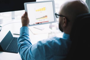 Mavi gömlekli bulanık bir adamın bilgisayar masasında elinde aletlerle oturup tabletten grafikler izlediği arka plan.