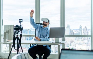 Yetişkin bir adam ofiste bilgisayarlı ve şehir manzaralı bir masada oturuyor ve tripodla kameranın önünde sanal gerçeklik gözlüklerini kullanıyor.