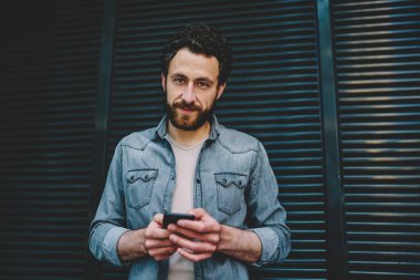 Sakallı hippi blogcu 4G interneti kullanarak internet uygulamasında profili güncellerken kameraya bakarken portresi. Kot giyen genç adam cep telefonuyla sosyal ağlarda sohbet ediyor.