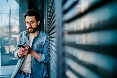 Yakışıklı beyaz erkek dışarıda akıllı telefon paylaşımı ile duruyor. Blogunda 4G bağlantısı ile yayınlanıyor. Günlük kıyafet giyen hippi adam sohbet etmek için cep telefonu kullanıyor.