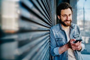 Neşeli sakallı hippi blogcu portresi. Duvarın yanında cep telefonu uygulaması yerleştirirken kameraya gülümsüyor. Sosyal ağlarda akıllı telefon cihazında sohbet eden genç adam.