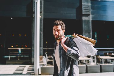 Havalı, sakallı genç bir adamın portresi kameraya bakarken arkasında modaya uygun eşyalar olan kağıt torbalar taşıyordu ve siyah cuma günü alışveriş merkezinin yanındaki şehir merkezinde yürüyordu.