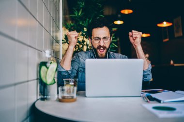 İnternet sitesinde dizüstü bilgisayarla yapılan kulaklıkla maç izlerken takım için tezahürat yapan mutlu sakallı genç adam. İnternet yarışmasında dizayn projesinin zaferini kutlayan neşeli hipster çocuk.