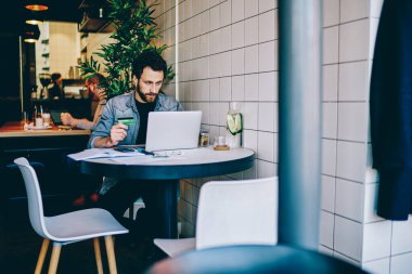 Düşünceli sakallı genç adam elinde kredi kartı tutuyor ve internet üzerinden internet üzerinden internet üzerinden para aktarıyor. Kafede oturan dizüstü bilgisayardaki kablosuz internet bağlantısını kullanıyor.